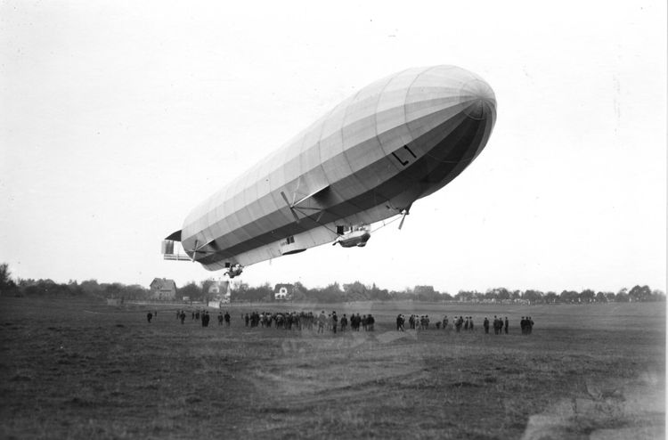 Die historische Aufnahme zeigt ein Luftschiff über einem Feld. Darunter sind mehrere Personen zu erkennen.