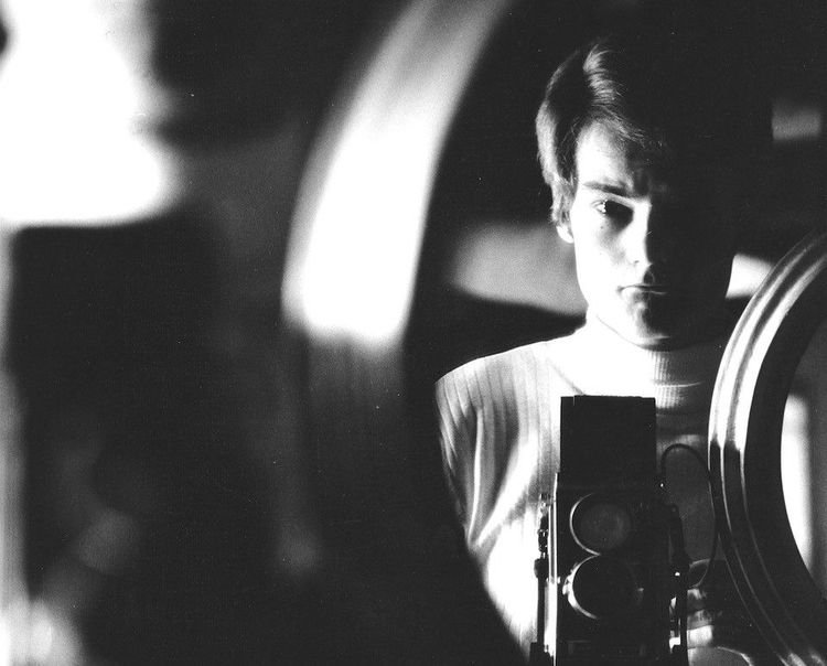 Die schwarz-weiß Fotografie zeigt das aufgenommene Selbstporträt eines jungen Mannes durch einen Spiegel. Der Mann trägt einen weißen Rollkragenpullover und hält eine Kamera vor der Brust.