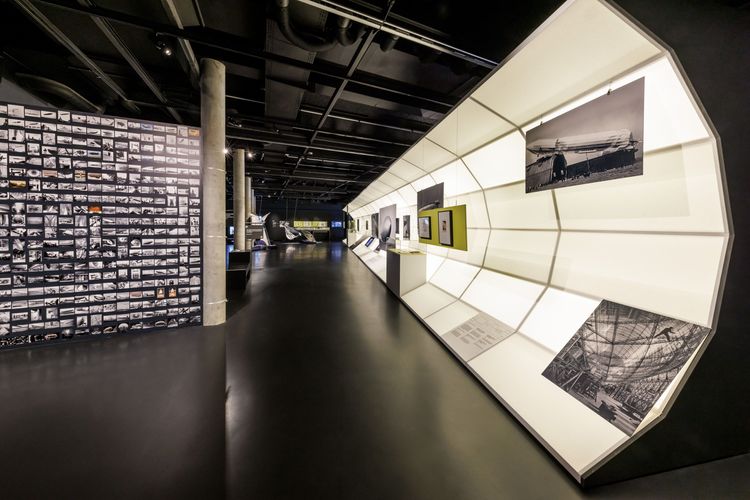 Eine dunkle Raumansicht mit einer halbrunden leuchtenden Wand, an der Fotos hängen, davor Personen