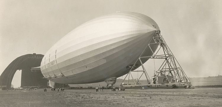 Das Luftschiff USS Akron, das vor einer Luftschiffhalle steht. Die Spitze wird mit einem Gerüst gestützt. 