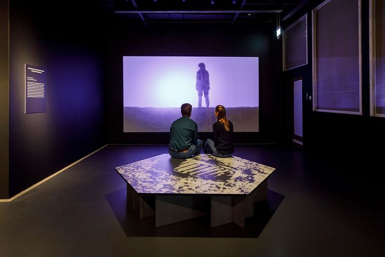 Die Ausstellungsansicht zeigt eine eckige Sitzbank auf der zwei Personen sitzen. Beide schauen auf eine Leinwand, die eine Person im Raumanzug zeigt. 