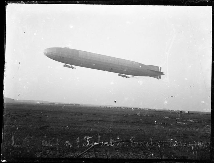 Schwarz-Weiß-Foto des LZ 54 L 19 in der Luft über einem Feld. Im Hintergrund sieht man flache Gebäude und eine große Menschenmenge. Auf einer schlecht lesbaren Inschrift ist „Erste Fahrt“ erkennbar. 