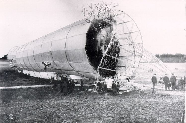 Schwarz-weiße Fotografie von LZ 2, kaputt auf einer Wiese. Die Bugspitze ist als Gerippe rechts vorne im Bild sichtbar, das schlauchartige Schiff dominiert die linke Bildhälfte. Unter dem Zeppelin stehen die Menschen.  