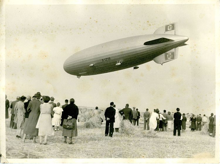 Schwarz-Weiß-Foto des LZ 130 „Graf Zeppelin“ über Viehtach im Bayrischen Wald. Unterhalb des Luftschiffs auf einem abgemähten Feld stehen zahlreiche Schaulustige zwischen einigen Heuballen.  