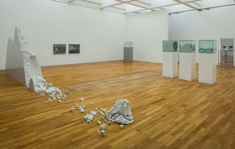 Die Ausstellungsansicht zeigt einen nachgebauten Eisberg, von dem Stücke abfallen. Außerdem stehen mehrere Podeste im Raum, auf denen sich gefüllte Glaskästen befinden. 