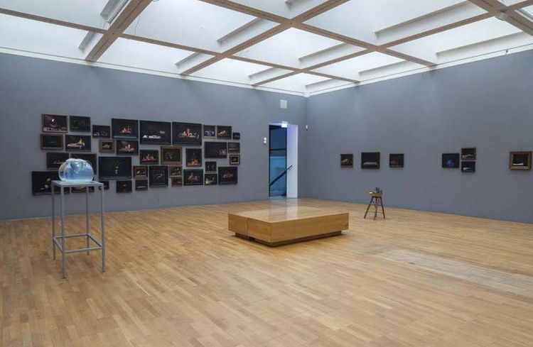 Die Ausstellungsansicht zeigt zwei graue Wände, an denen mehrere Bilderrahmen hängen. Auf einem Podest stehen zwei runde Glasobjekte.