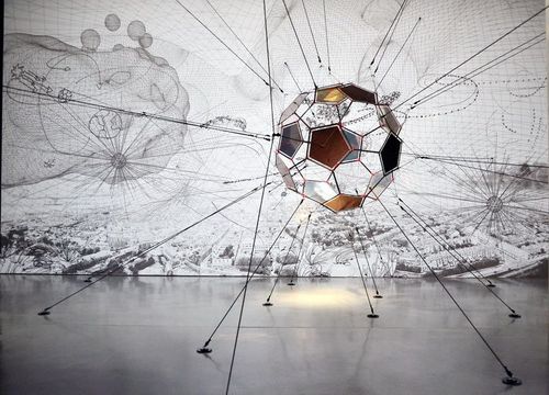 Eine große, runde Installation, von der Seile an verschiedene Punkte der Decke und des Bodens verlaufen befindet sich mittig im Bild. Dahinter an der Wand befindet sich ein großes, beleuchtetes Bild.