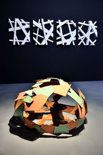 Eine rundliche Installation, die aus mehreren verbundenen Platten besteht, liegt auf dem Boden. Dahinter an der Wand hängen vier Bilder.