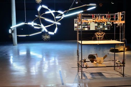 Rechts im Bild steht ein Regal auf dem sich verschiedene Objekte befinden. Dahinter hängt eine große, leuchtende Installation von der Decke.