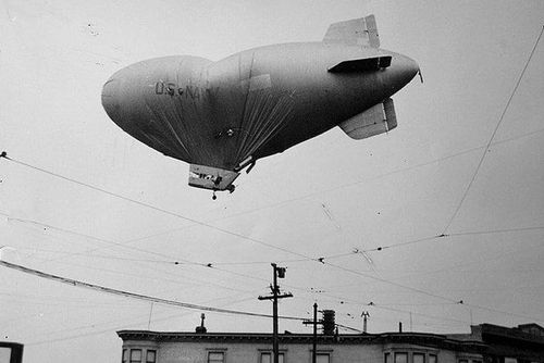 Die historische Aufnahme zeigt ein Prallluftschiff am Himmel, dessen Hülle beschädigt ist und einknickt.