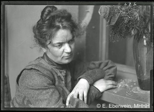 Die historische Aufnahme zeigt das Portrait einer sitzenden, älteren Frau mit hochgesteckten Haaren. Die Frau blickt nachdenklich und mit gesenktem Kopf rechts an der Kamera vorbei. 