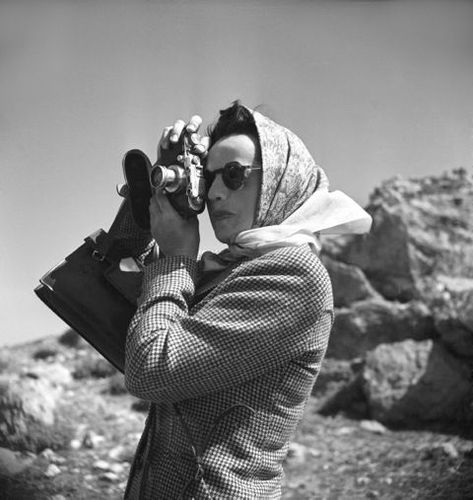 Die schwarz-weiß Fotografie zeigt eine Frau, die gerade ein Foto zu schießen scheint. Sie trägt eine Sonnenbrille und ein Tuch um den Kopf. Mit beiden Händen hält sie eine Kamera und blickt durch den Sucher. 