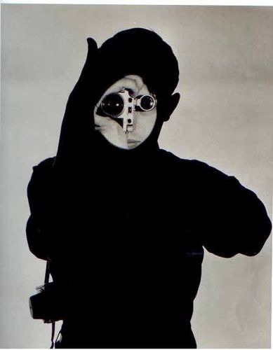 Die schwarz-weiß Fotografie zeigt die schwarze Silhouette eines Mannes, der eine Kamera vor dem Gesicht hält. Nur ein kleinerer Kreis über dem Gesicht ist beleuchtet und zeigt die Kamera vor den Augen, der Rest des Mannes bildet einen schwarzen Schatten.