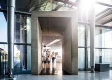 Blick in einen Ausstellungsraum mit Glasfassade. In der Mitte steht ein großer, rechteckiger Eingang aus Beton. Eine Frau und ein Kind gehen in den hell beleuchteten Raum dahinter.