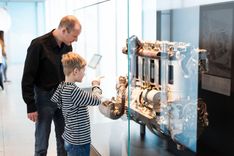 Ein Junge und ein Mann stehen vor einer großen Glasvitrine, in der sich ein großes, technisches Objekt befindet. Dabei könnte es sich um eine Art Motor handeln.
