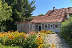 Außeansicht des Schauhaus vom Garten aus gesehen. Blühende Blumensträucher sind im Vordergrund, Keis führt bis zur Rückseite des Hauses. 