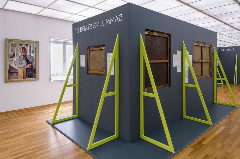 Ein heller Ausstellungsraum mit einer grauen Wandkonstruktion in der Mitte, an der mehrere Gemälderückseiten präsentiert sind. Die Konstruktion wird von leuchtend grünen Stützen gehalten. Links ist ein farbiges Gemälde an der Wand zu sehen.