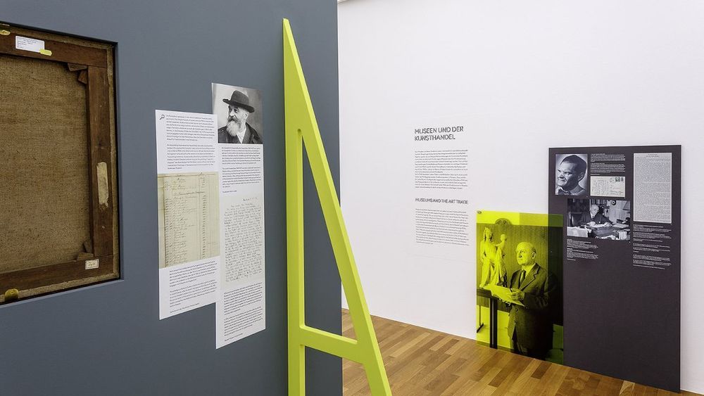 Ein Ausstellungsraum mit mehreren Texttafeln, Fotos und einer gelben, dreieckigen Konstruktion in der Mitte. An der linken Wand hängt die Rückseite eines gerahmten Bildes. Im Hintergrund sind Porträts und Informationen zu Personen ausgestellt.