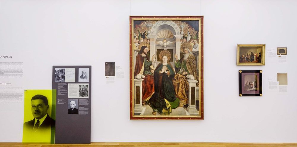 Ein Museumsraum mit weißer Wand, an der mehrere Gemälde in unterschiedlichen Größen hängen. In der Mitte befindet sich ein großes religiöses Gemälde. Links daneben sind Infotafeln und Fotos von einer Person zu sehen.
