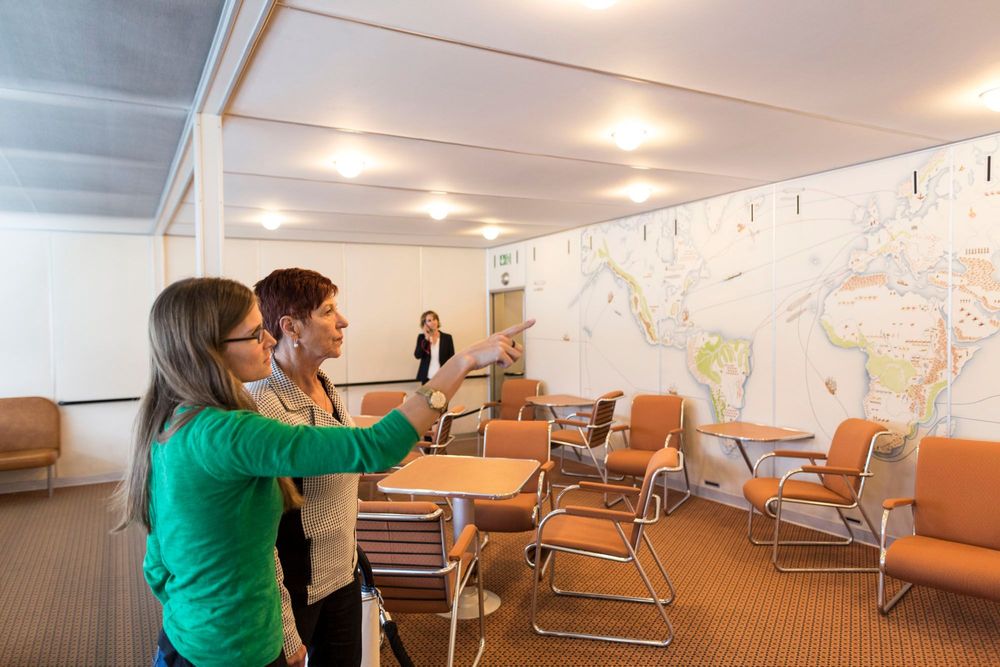 Zwei Frauen stehen in einem Raum mit braunen Sesseln und Tischen. Eine Frau zeigt auf eine große Weltkarte an der Wand. Im Hintergrund ist eine weitere Person zu sehen.