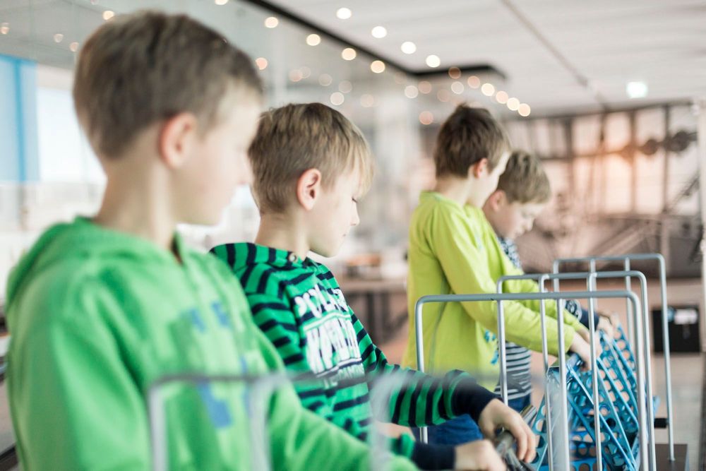 Vier Jungen stehen nebeneinander vor einer Mitmachstation. Zwischen Stäben befindet sich eine Art kleines Gerüst, welches die Jungen versuchen anzuheben.