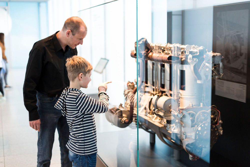 Ein Junge und ein Mann stehen vor einer großen Glasvitrine, in der sich ein großes, technisches Objekt befindet. Dabei könnte es sich um eine Art Motor handeln.