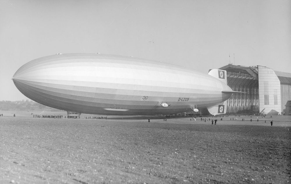Historische Schwarz-Weiß-Aufnahme eines Luftschiffs, das sich auf einem flachen Feld vor einer Luftschiffhalle befindet.