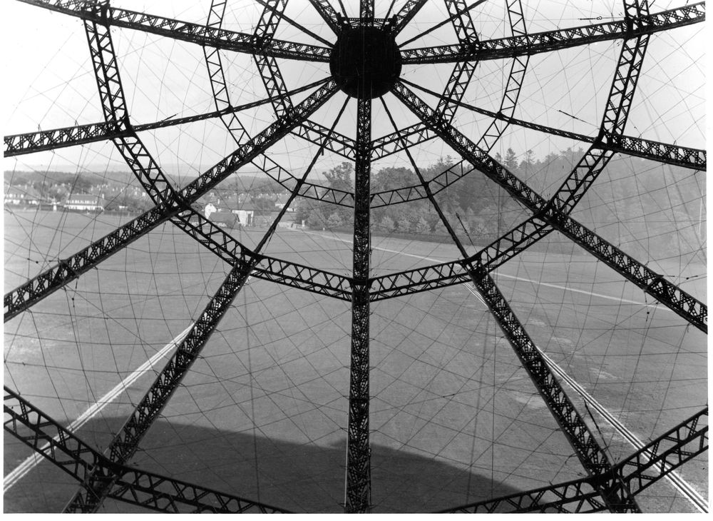 Schwarz-Weiß-Fotografie einer kreisförmigen Metallkonstruktion, von der Verstrebungen strahlenförmig ausgehen. Durch das Gitterwerk blickt man auf den Boden eines Feldes.