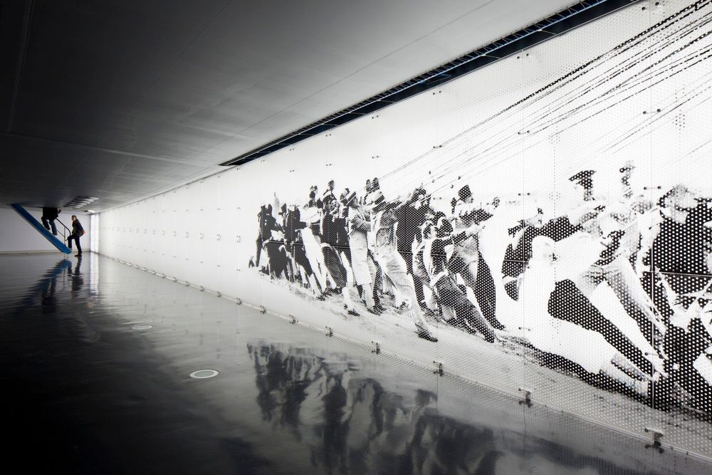 An einer hell leuchtenden weißen Wand ist ein schwarz-weiß Foto einer Haltemannschaft abgebildet. Die Männer auf dem Bild sind in gebeugter Haltung und halten an verschiedenen Seilen fest.