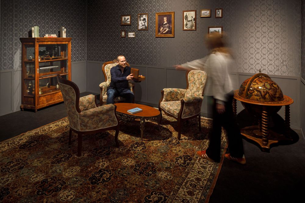 Zwei Personen spielen im Escape Room vom Zeppelin Museum. Das ist ein Wohnzimmer mit alten Möbeln.