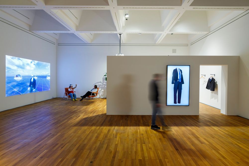 Die Ausstellungsansicht zeigt zwei Personen die auf Sesseln sitzen. Eine der beiden Personen trägt eine VR-Brille und hält Kontroller in den Händen. Im Vordergrund läuft eine Person an einer Wand vorbei, an der ein großer Bildschirm hängt.