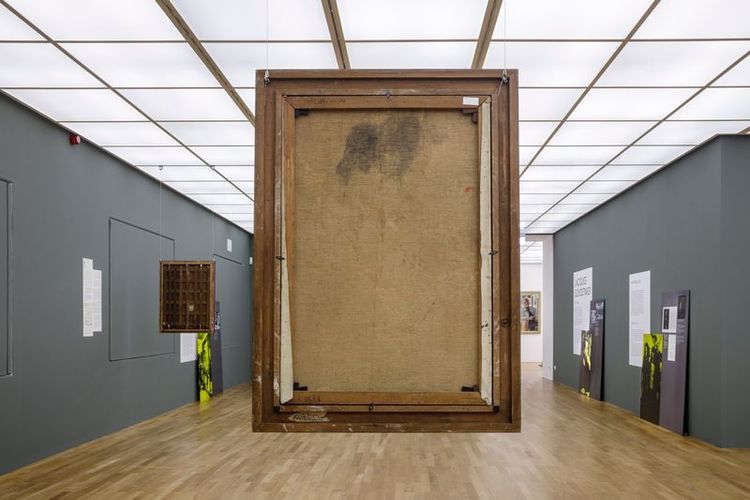 Blick in einen Ausstellungsraum mit einem großen, umgedrehten Gemälde im Mittelpunkt, sodass die Rückseite sichtbar ist. An den Wänden hängen Infotafeln